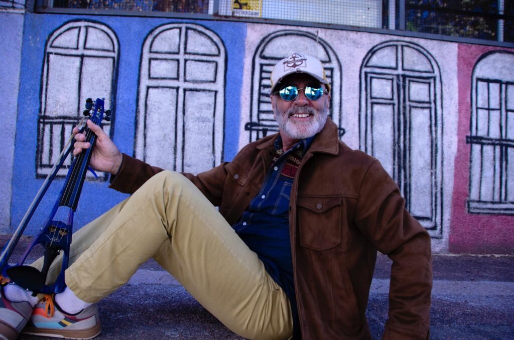 Edison Mouriño sonriendo con su violín eléctrico azul frente a un mural pintado