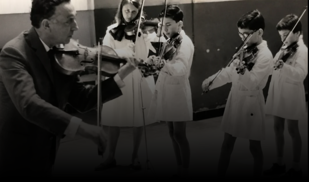Clase de violín en la época de formación de Edison Mouriño: fotografía en blanco y negro de un maestro tocando junto a alumnos con túnica escolar
